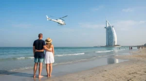 Two visitors on Jumeirah beach watch a white helicopter fly past over the sea, with the Burj Al Arab beyond