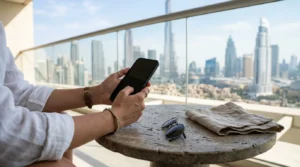 Traveller's hands holding a phone over a hotel balcony table, with the Dubai skyline softly blurred behind