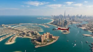 A red and white helicopter flies over the Palm Jumeirah and Atlantis resort toward Dubai's distant skyline.